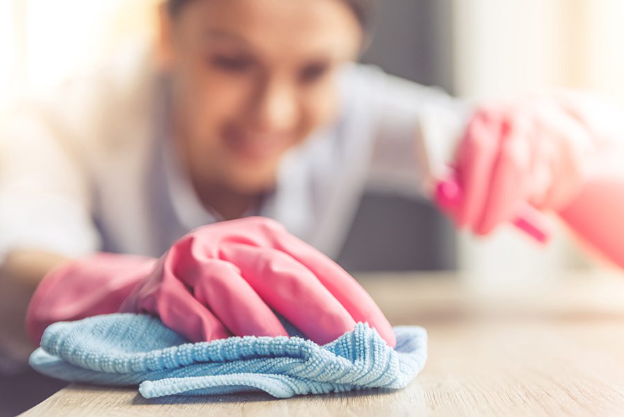 Woman in protective gloves is smiling and wiping dust using a spray and a duster while cleaning her house, close-up